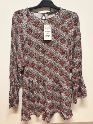 Mönstrad röd och vit playsuit från Zara - Playsuit från Zara storlek M med röd och vit blommig print. Långa ärmar med volangdetalj vid ärmslut och öppning i ryggen med knapp. Tillverkad i ett lätt och flowigt tyg som ger en avslappnad känsla. Perfekt för dig som gillar färg och mönster.