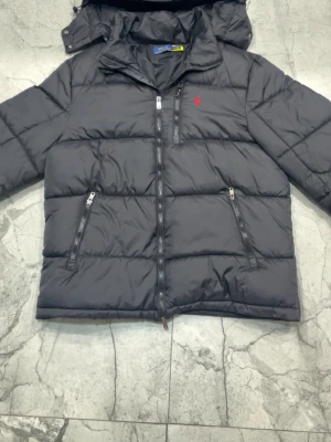 Svart pufferjacka från Polo Ralph Lauren - Svart pufferjacka från Polo Ralph Lauren med röd logga på bröstet. Jackan har två dragkedjeförsedda fickor framtill och hög krage. Materialet är glansigt och quiltat, perfekt för kalla vinterdagar. Klassisk och stilren design med robust dragkedja.