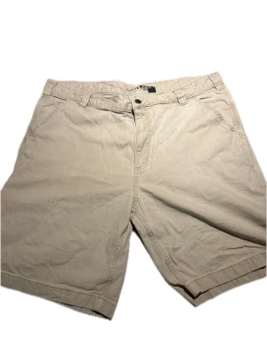 Beige shorts från Carhartt - Beiga shorts från Carhartt med klassisk passform och flera fickor, inklusive bakfickor och en sidoficka. Tillverkade i slitstark bomull med bälteshällor och knappstängning. Perfekta för en avslappnad stil.