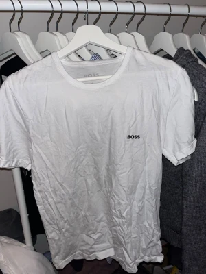 Vit t-shirt från BOSS - Vit t-shirt från BOSS med klassisk rund halsringning och diskret BOSS-logga på bröstet. Enkel och stilren design som passar till det mesta. Tillverkad i mjuk bomull och har korta ärmar.