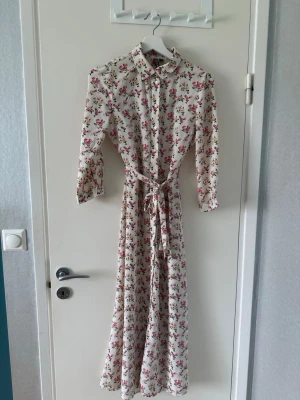 Långklänning - Långklänning i vit bas med blommigt mönster i rosa och grönt. Klänningen har lång ärm, krage och knappar framtill samt ett matchande knytband i midjan. Perfekt för sommarens alla tillfällen.