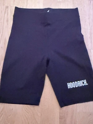 Svarta cykelshorts från Hoodrich - Svarta cykelshorts från Hoodrich med vit logga tryckt på ena benet. Tight passform och stretchigt material som passar perfekt till streetwear eller träning. Snygg minimalistisk design utan fickor eller extra detaljer.