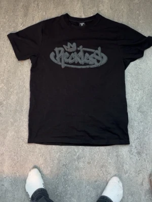 Svart t-shirt från FSBN med tryck - Cool svart t-shirt från FSBN med stort grått 'Reckless' tryck på bröstet och regular fit. Perfekt för dig som gillar streetstyle och grafiska prints. Rund halsringning och korta ärmar. Materialet är mjuk bomull som känns skönt mot huden.