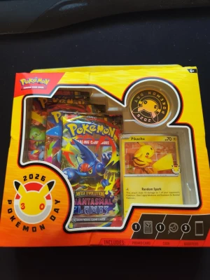 Pokémon Day 2026 samlarkortbox - Säljer en Pokémon Day 2026 samlarkortbox med flera booster packs och en Pikachu promo-kort. Boxen är gul med färgglada detaljer och innehåller samlarkort från Mega Evolution Phantasmal Flames-serien. Perfekt för Pokémon-entusiaster.