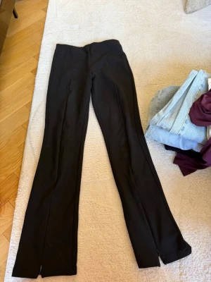 Svarta kostymbyxor med slits - Snygga svarta kostymbyxor med raka ben och diskreta slitsar vid bensluten. Byxorna har en klassisk passform och är tillverkade i ett mjukt, stretchigt tyg som ger en skön känsla. Perfekta för en clean och stilren look.