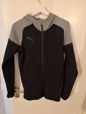 Svart och grå hoodie från Puma - Snygg hoodie från Puma i svart med gråa axlar och huva. Dragkedja framtill och två sidofickor. Puma-logga på bröstet. Perfekt för chill eller träning. Tillverkad i mjukt och bekvämt material som känns skönt mot huden.