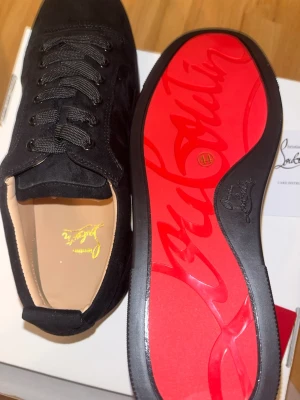 Svarta sneakers från Christian Louboutin - Snygga svarta sneakers från Christian Louboutin i mocka med klassisk röd sula och guldfärgad logga på innersulan. Skorna har svarta skosnören och låg profil som ger en clean och exklusiv look. Perfekta för dig som gillar lyxiga detaljer och streetstyle. Grish passar i kostym och som daglig stil. Pris kan diskuteras 