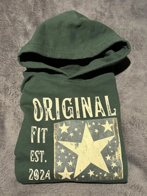 Mörkgrön hoodie med stjärntryck - Snygg mörkgrön hoodie med stor beige stjärna och texten 'ORIGINAL FIT EST. 2024' på bröstet. Hoodien har huva och en klassisk känguruficka framtill. Perfekt för dig som gillar streetwear och vill sticka ut med ett coolt tryck.