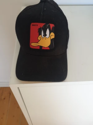 Svart keps med Daffy Duck från Capslab - Säljer en svart truckerkeps från Capslab med ett stort Daffy Duck-märke framtill. Kepsen har mesh på sidorna och justerbar snapback baktill. Perfekt för dig som gillar Looney Tunes och vill ha en cool accessoar med karaktär.