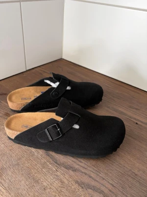 Svarta Birkenstock Boston tofflor - Säljer ett par svarta Birkenstock Boston tofflor i mocka med klassisk design och justerbart spänne. De har en bekväm innersula i kork och gummisula undertill. Perfekta för en avslappnad och stilren look.