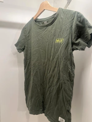 Grön t-shirt med tryck från lager 157 - Mörkgrön t-shirt från lager 157med korta ärmar och rund hals. På bröstet finns ett gult tryck med texten 'HELLO!' och på ryggen står det 'HOW U DOIN?'. Skön och enkel stil, perfekt till jeans eller shorts.
