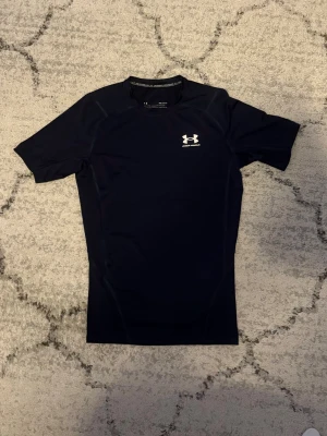 Kompressionströja Under Armour - Svart kortärmad kompressionströja från Under Armour med vit logga på bröstet. Tillverkad i ett stretchigt och fukttransporterande material som passar perfekt för träning och sportaktiviteter. Tight passform och rund halsringning.