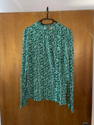Grön blommig blus från Holly & Whyte - Grön blus med vitt blommönster från Holly & Whyte by Lindex. Blusen har långärmad design, rund halsringning och knapp i nacken. Tunt och luftigt material som ger en skön känsla. Perfekt för dig som gillar en avslappnad men ändå trendig stil.