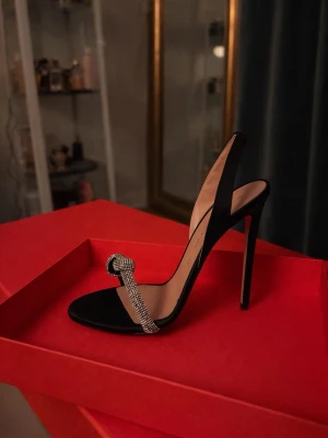 Svarta pumps med strassdetalj - Identita Kimber satin Heel - Säljer ett par svarta pumps (strl 39)  med öppen häl och smal hög klack. Skorna har en elegant rem framtill dekorerad med gnistrande strass och en snygg knutdetalj. Perfekta för dig som vill sticka ut med en lyxig och trendig look. Skorna är helt nya se bild endast provade inomhus.  Ovandel: Satin * Rem med knut prydd med kristaller * Insida: Äkta läder * Passform: Normal  * Halkfri sula * Klacktyp: Stilettklack |12mm  * Tå: Rund