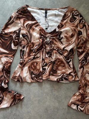 Brun swirlmönstrad topp med snörning - Trendig långärmad topp med swirlmönster i bruna och beige toner. Toppen har v-ringning, snörning framtill och utsvängda ärmslut. Perfekt för dig som gillar unika detaljer och en cool look.