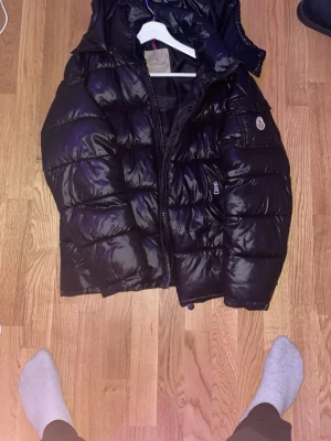Svart pufferjacka från Moncler - Säljer en svart, glansig pufferjacka från Moncler med huva och dragkedja framtill. Jackan har en klassisk quiltad design och Moncler-logga på ärmen. Materialet är syntet med dunfyllning för extra värme. Perfekt för kalla dagar och riktigt snygg streetstil.