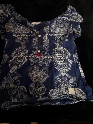 Blå paisleymönstrad blus Odd Molly - Snygg blå blus från Odd Molly med vitt paisleymönster och dekorativa knappar framtill. Blusen har korta ärmar och en liten rosett som detalj. Tillverkad i ett lätt och luftigt material, perfekt för en avslappnad stil.