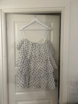 Vit prickig volangklänning - Söt vit kortklänning med svarta prickar och flera volanger. Klänningen har tunna axelband och en luftig, flowy passform som känns perfekt för varma dagar. Materialet är lätt och känns som polyester. Perfekt för dig som gillar lekfulla och trendiga plagg.