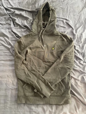 Olivgrön hoodie från Lyle & Scott - Snygg olivgrön hoodie från Lyle & Scott med klassisk gul logga på bröstet. Hoodien har dragsko i huvan, känguruficka framtill och ribbade muddar vid ärmslut och nederkant. Perfekt för en avslappnad och stilren look.