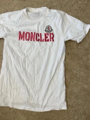 Vit Moncler t-shirt med röd logga - Säljer en vit t-shirt från Moncler med röd textlogga och klassisk Moncler-patch på bröstet. T-shirten har korta ärmar och en normal passform. Perfekt för dig som gillar stilrena och trendiga plagg med tydlig branding.