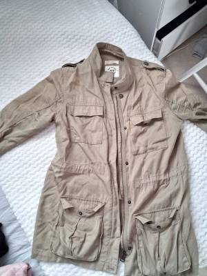 Beige vindjacka med tryck - Stilren grön vindjacka från Lava med coolt vitt tryck på ryggen: 'IS IT FRIDAY YET? #WEEKENDFOREVER'. Jackan har fyra stora fickor framtill, axelklaffar och dragkedja med tryckknappar. Perfekt för dig som gillar streetstyle och praktiska detaljer.Grön jacka i fint skick 💚  Säljer en snygg grön jacka som tyvärr inte används längre. Perfekt för både vardag och lite kallare dagar.  