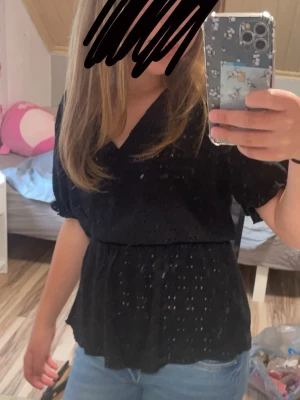 Svart topp från vero moda - Säljer en svart omlottblus med korta puffärmar och fina spetsdetaljer. Blusen har v-ringning och resår i midjan för en snygg passform. Perfekt till jeans eller kjol för en trendig look💗Kolla in min profil för fler fina och trendiga plagg!