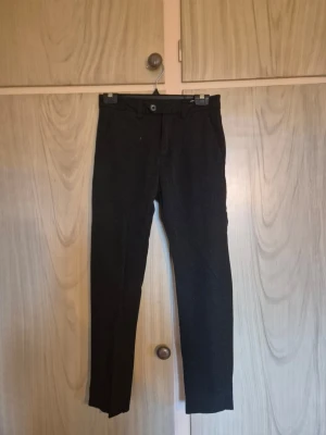 Svarta kostymbyxor med smal passform - Snygga svarta kostymbyxor med smal passform och klassisk design. Byxorna har bälteshällor, knapp och dragkedja framtill samt två bakfickor med knapp. Perfekta för dig som vill ha en stilren look. Materialet ser ut att vara mjukt och följsamt.    Alla kläder rollas innan dom skickas hem då det finns katt i hemmet🐈 Jag stryker och tvättar INTE innan jag skickar det får du stå för själv så det blir som du vill ha det🤗