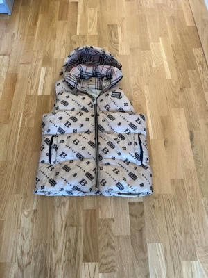 Beige dunväst från Burberry - Snygg beige dunväst från Burberry med ikoniskt rutigt foder och svart logotypmönster över hela västen. Västen har dragkedja framtill, två fickor med dragkedja och en stor huva. Perfekt för lager på lager och streetstyle.