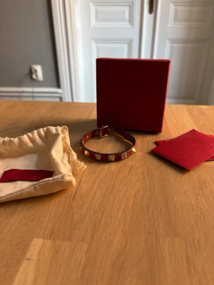 Rött Valentino armband med nitar - Säljer ett snyggt armband från Valentino i rött läder med guldiga pyramidformade nitar runt om. Armbandet har justerbart spänne och kommer med originalförpackning och dustbag. Perfekt accessoar för att lyfta din outfit.