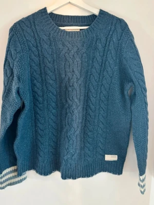 Blå kabelstickad tröja Odd Molly - Snygg blå kabelstickad tröja från Odd Molly med rund halsringning och ribbade muddar. Tröjan har ett klassiskt flätmönster framtill och diskreta vita och blå ränder på ena ärmens mudd. 