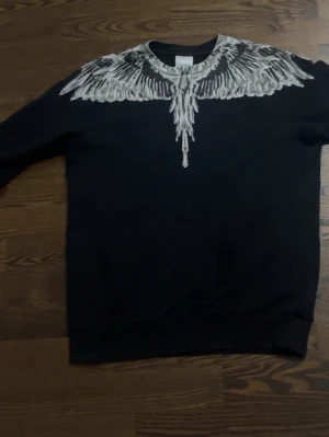 Marcelo burlon tröja äkta - Säljer en svart sweatshirt från Marcelo Burlon med ikoniskt vitt fjädermönster runt halsen och över axlarna. Tröjan har rund halsringning och är tillverkad i mjukt bomullsmaterial. Perfekt för dig som vill sticka ut med en unik design.