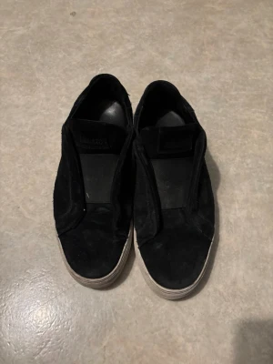 Svarta slip-on sneakers från Vagabond - Stilrena svarta slip-on sneakers från Arigato med ovandel i mocka och vit sula. Skorna har en enkel och clean design utan snörning, vilket gör dem lätta att ta på och av. Perfekta för en avslappnad och trendig look.