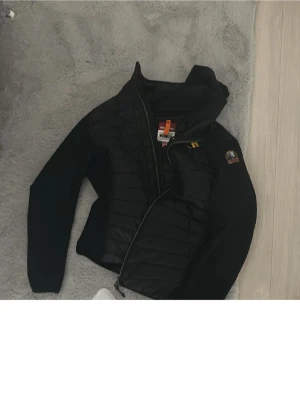 Svart hybridjacka från Parajumpers - Säljer en svart hybridjacka från Parajumpers med quiltad front och mjuka ärmar. Jackan har huva, dragkedja och coola detaljer som logopatch på ärmen och orange flärp vid halsen. Perfekt för dig som vill ha en stilren och funktionell jacka. Använd några gånger är som helt ny.