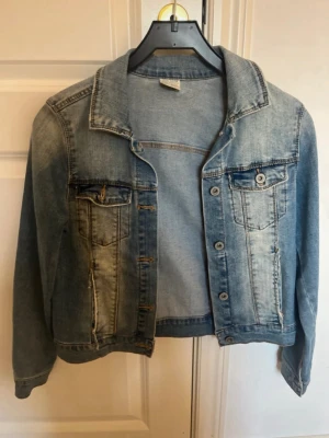  blå jeansjacka - Snygg blå jeansjacka med krage och knäppning framtill. Jackan har två bröstfickor med lock och knapp samt två sidfickor. Färgen är ljusblå med slitningar för en trendig look. Perfekt att slänga över en hoodie eller t-shirt.