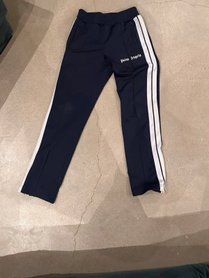 Palm Angels marinblå mjukisbyxor - Palm Angels mjukisbyxor i marinblått med vita sidoränder och logga på vänster ben. Byxorna har resår i midjan och raka ben. Perfekta för en avslappnad och trendig streetwear-look.