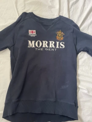 Mörkblå sweatshirt från Morris - Mörkblå sweatshirt från Morris med broderad logga och texten 'MORRIS THE GENT' på bröstet. Detaljer som brittisk flagga och emblem på framsidan samt siffran 68 i vitt på ena ärmen. Skön passform och ribbade muddar. Perfekt för en sportig och stilren look.