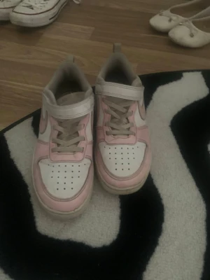 Nike sneakers i vitt och rosa - Nike sneakers i low-modell med vit bas och ljusrosa detaljer. Skorna har klassisk snörning kombinerat med kardborreband och är tillverkade i syntetmaterial och textil. Perfekta för en sportig och trendig look.