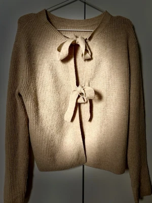 Beige stickad kofta  - Jättefin mysig beige stickad kofta med två knytband framtill. Koftan har ribbstickad struktur, rund halsringning och långa ärmar. 