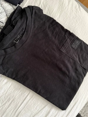 Svart t-shirt från Etirel med ficka - Svart t-shirt från Etirel i bomull med klassisk passform. T-shirten har en bröstficka med en liten patchdetalj och rund halsringning. Perfekt basic-plagg att ha i garderoben, stor i storlek skulle jag säga!