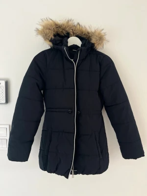 Svart dunjacka med fuskpäls - Snygg svart dunjacka med huva och fuskpälsdetalj. Jackan har dragkedja framtill, två stora fickor och justerbar midja. Perfekt för kalla vinterdagar och ger en stilren look.