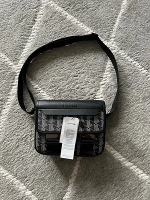 Lacoste axelväska  - Snygg svart axelväska från Lacoste med justerbar rem och lock. Väskan har ett grafiskt mönster i vitt med Lacoste-logga på locket och detaljer i svart konstläder. Perfekt för att bära dina viktigaste grejer med stil.
