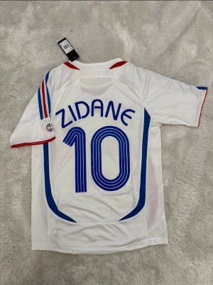 Frankrike Zidane matchtröja Adidas - Säljer en vit matchtröja från Frankrikes landslag med Zidane och nummer 10 på ryggen. Tröjan har blå och röda detaljer, broderad landslagslogga och Adidas-logga på bröstet. Tillverkad i lätt och ventilerande material, perfekt för fotboll eller samlare.