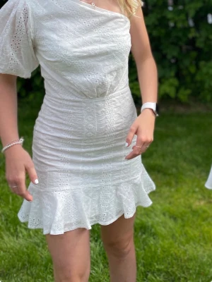 Vit one shoulder klänning med volang - Superfin vit kortklänning med one shoulder och volangkant nertill. Klänningen har broderade detaljer och är tillverkad i ett lätt bomullstyg. Perfekt för sommarens alla tillfällen!