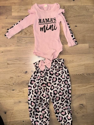 Rosa bodyset med leopardmönster - Sött set med rosa ribbad body med texten 'MAMA'S mini' och volangdetaljer på axlarna. Tillhörande byxor i rosa med svart och brun leopardmönster samt matchande rosett. Perfekt för en trendig liten tjej.