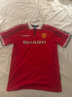 Manchester United tröja Beckham #7 - Klassisk röd Manchester United matchtröja från Umbro med SHARP som sponsor, klubbmärke på bröstet och svartvita detaljer på krage och ärmslut. På ryggen står det Beckham och nummer 7. Perfekt för fotbollsfans! Står att storleken är XL men är som M