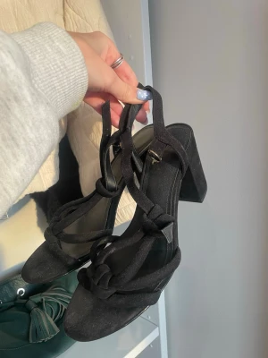 Svarta klackar med knutdetaljer - Säljer ett par svarta klackar med chunky klack och snygga knutdetaljer på remmarna. Skorna har öppen tå och är gjorda i mockaliknande material. Perfekta för att lyfta vilken outfit som helst med sin trendiga design.