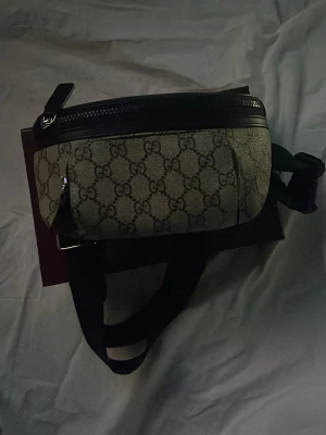 gg beige magväska med GG-mönster - Snygg magväska från gg i beige canvas med klassiskt GG-mönster och svarta läderdetaljer. Väskan har justerbart svart band och dragkedja upptill samt en ficka på baksidan. Perfekt accessoar för att lyfta din outfit.