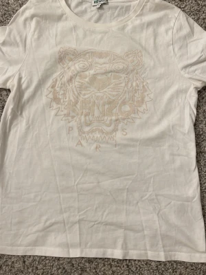 Vit Kenzo t-shirt med tigertryck - Säljer en vit t-shirt från Kenzo med det ikoniska tigertrycket och texten 'Paris' framtill. T-shirten har rund hals och korta ärmar. Perfekt för dig som gillar streetwear och vill ha något stilrent men ändå med attityd.