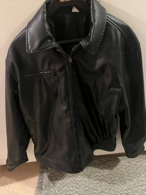 Svart klassisk skinnjacka - Säljer en svart jacka  dragkedja. Jackan har bred krage och resår i midjan för en snygg passform. Perfekt för dig som gillar tidlös och stilren look.
