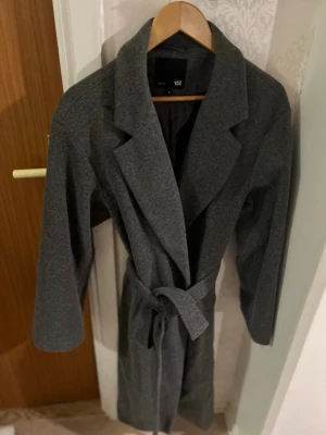 Mörkgrå kappa med knytskärp - Stilren mörkgrå kappa från 157 med klassisk krage och knytskärp i midjan. Kappan har en rak passform och är tillverkad i ett mjukt tyg som ger en snygg siluett. Perfekt för dig som vill ha en enkel men trendig look.